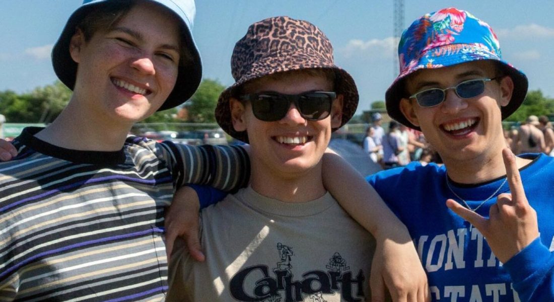 Tre unge venner i farverige bucket hats smiler og hygger sig til Skive Festival på Strandtangen i solskin.