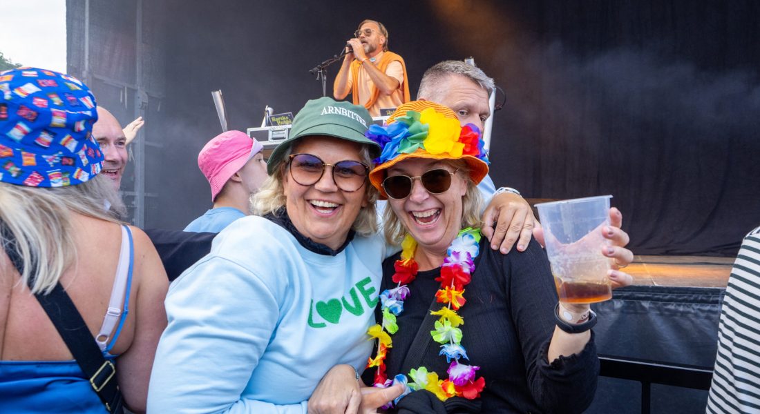 To smilende kvinder med farverige festivalhatte og blomsterkrans foran en scene til Skive Festival på Strandtangen.