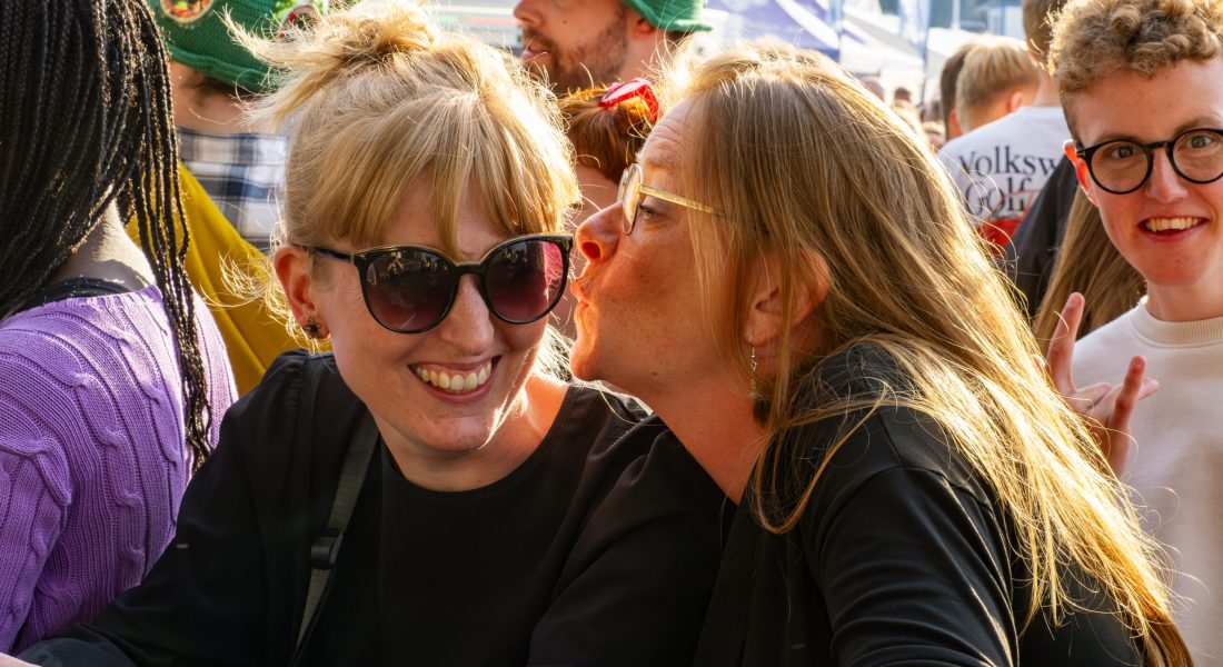 To glade kvinder i publikum på Skive Festival, hvor den ene giver den anden et kys på kinden i sommerens solskin.