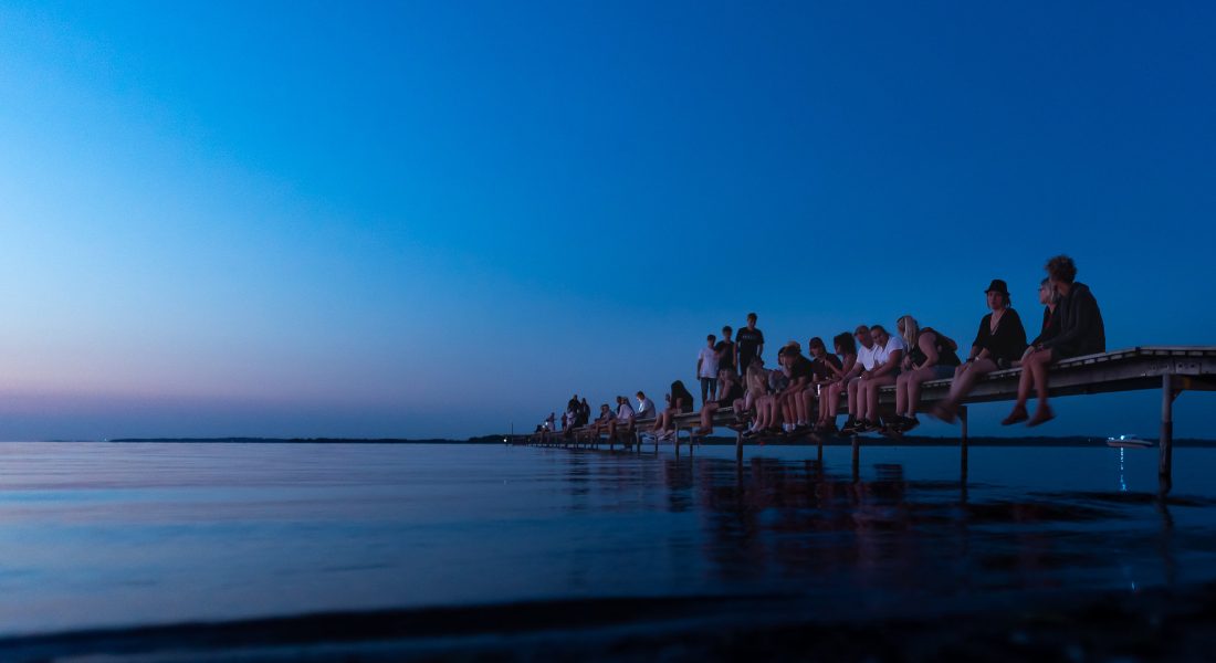 En gruppe unge mennesker sidder på en badebro ved vandet i skumringen under Skive Festival på Strandtangen.