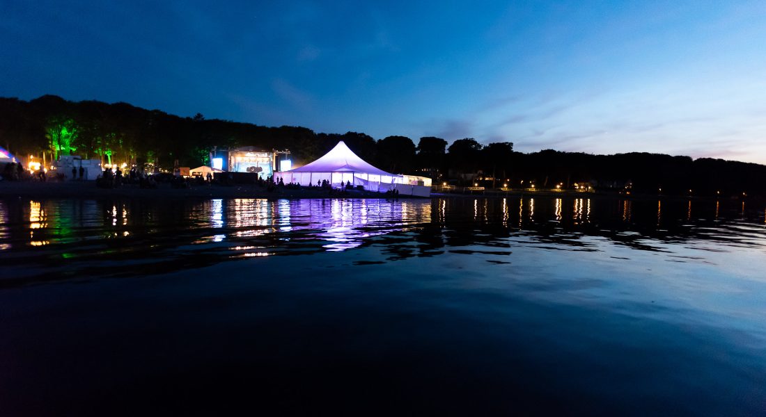 Oplyst hvidt festivaltelt ved vandet om aftenen til Skive Festival på Strandtangen med spejlinger i vandoverfladen.